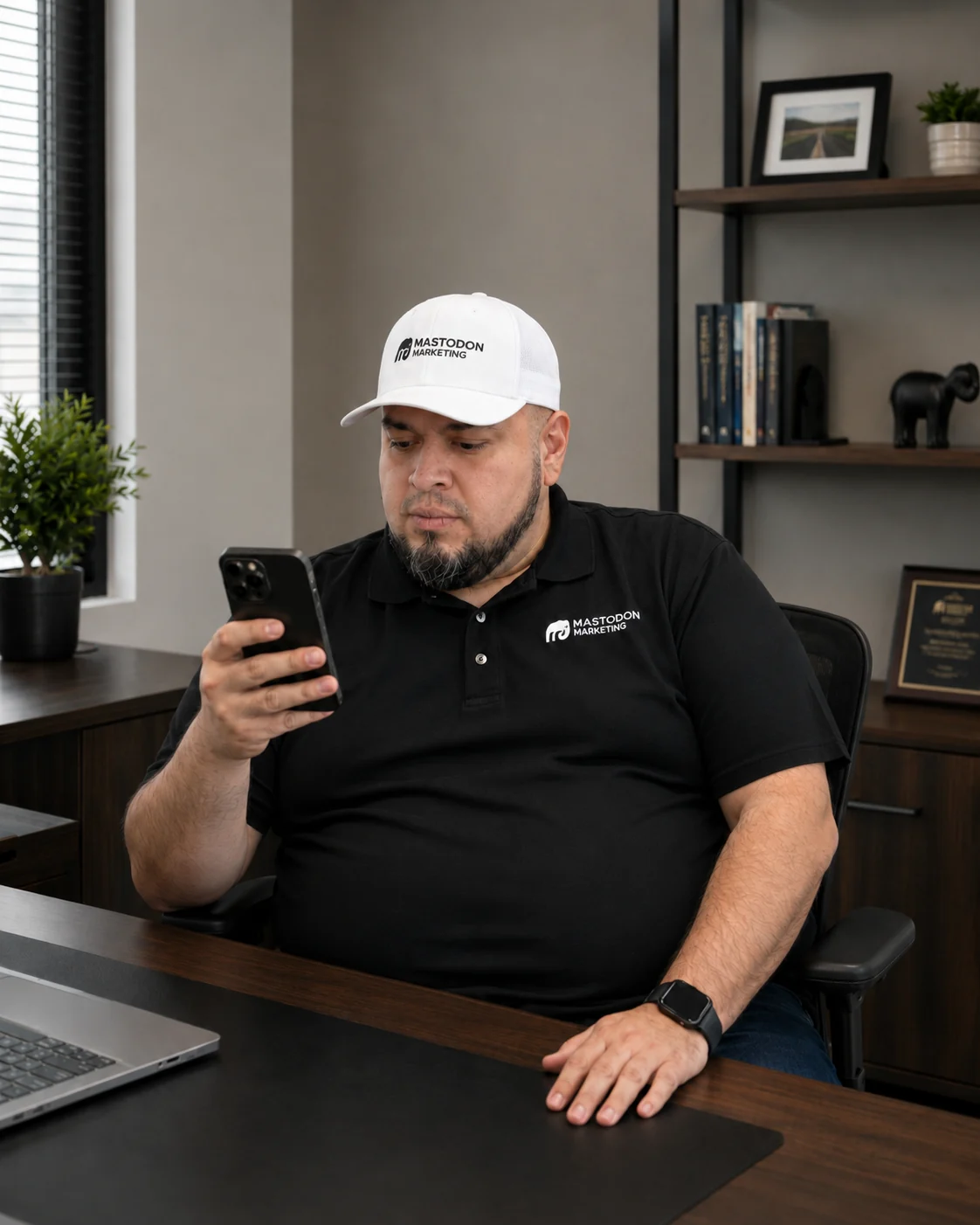 Alex Rocha at his desk reviewing client notifications on his phone at Mastodon Marketing's Pearland Texas office wearing a Mastodon Marketing branded polo and cap